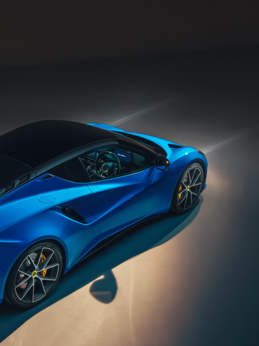 Angolo posteriore destro rialzato di una Lotus Emira. blu Gli splendenti fasci dei fari vengono proiettati visibilmente dalla parte anteriore dell'auto. | 压缩-I4.jpg