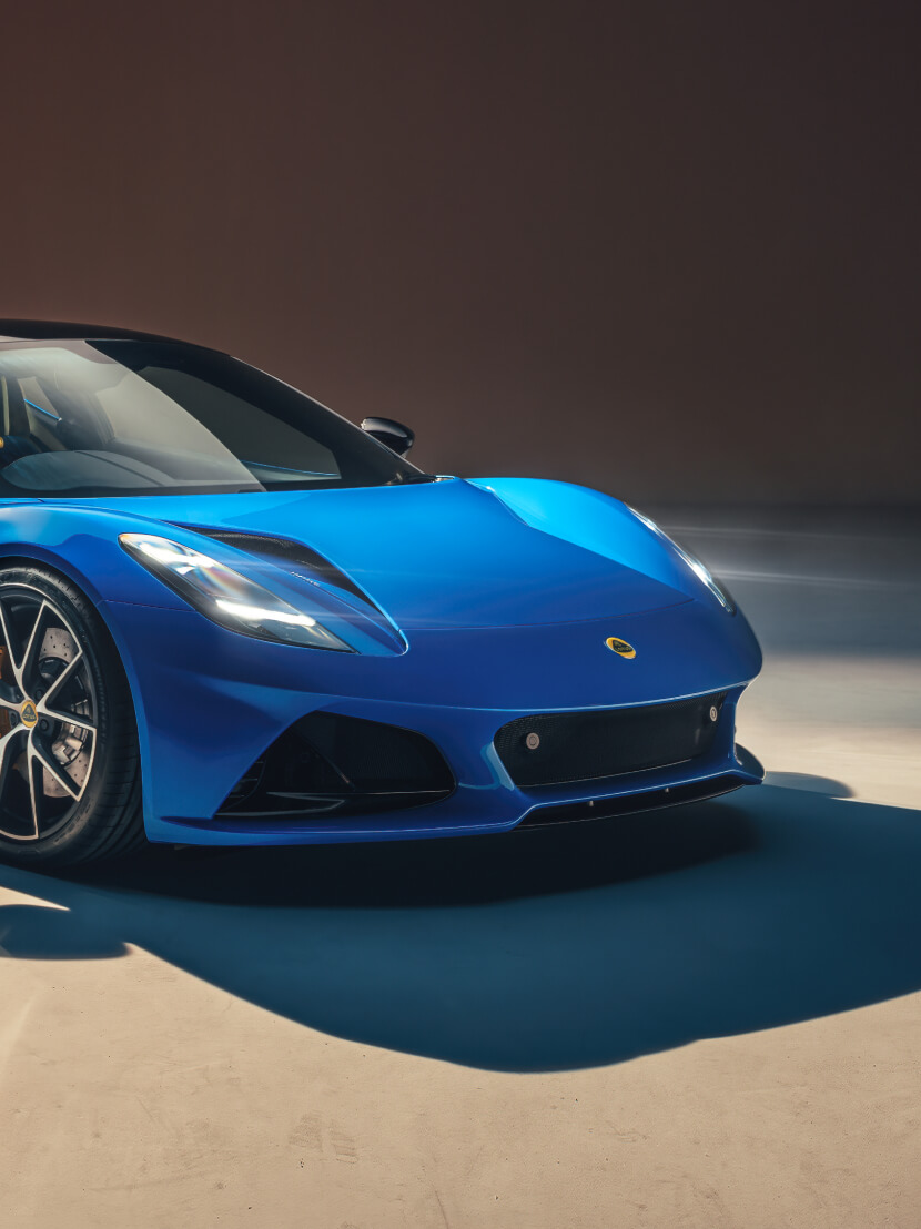 Angle de face avant d’une Lotus Emira bleue dans un environnement fortement éclairé. L’éclairage crée de fortes ombres autour du nez de la voiture. | 压缩-v6.jpg