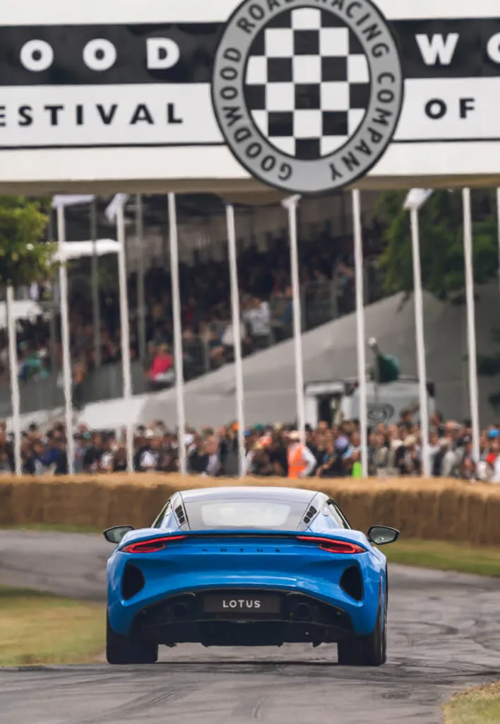 Rear shot of a blue Lotus Emira driving under a Goodwood Festival of Speed banner