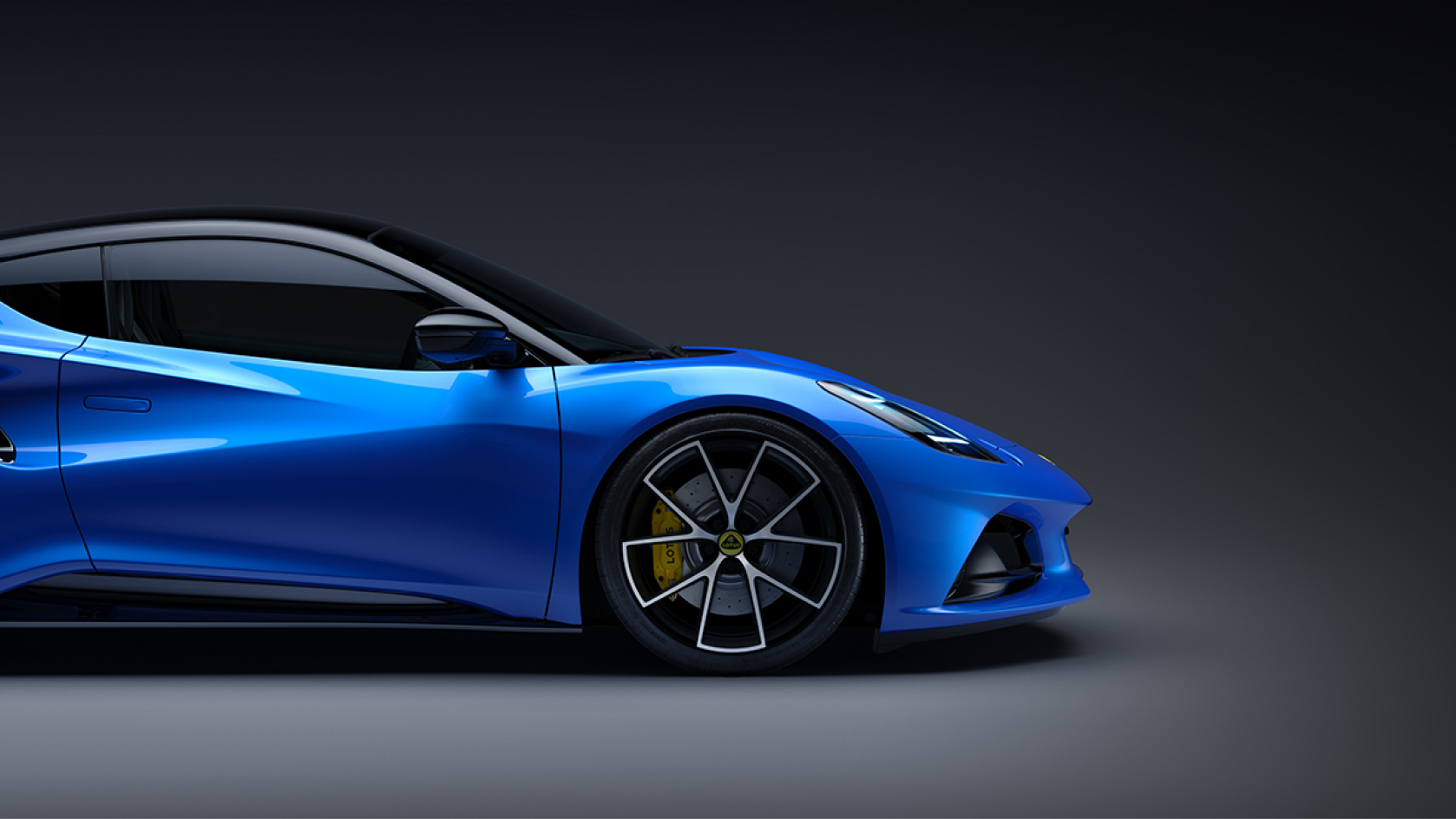 Profil der Vorderseite eines blauen Lotus Emira in einer abstrakten grauen Umgebung. Die gelben Bremssättel und das gelbe Lotus-Rademblem sind sichtbar. | 0701 Performance.jpg
