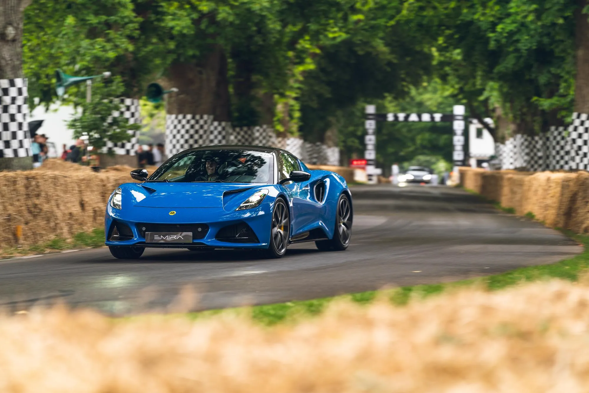 Front angle of a blue Lotus Emira driving along a tree-lined road at the Goodwood Festival of Speed